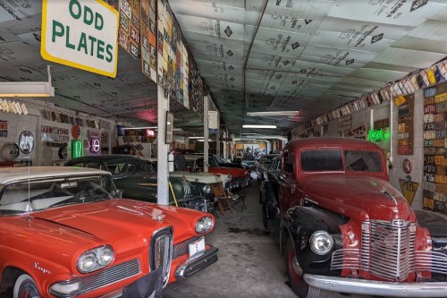 A vintage car garage filled with classic cars, neon signs, and an assortment of license plates on the walls.