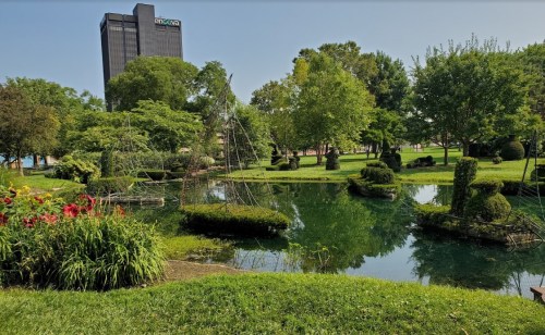 Lush green park with a pond, trees, and decorative shrubs, set against a backdrop of a tall building.
