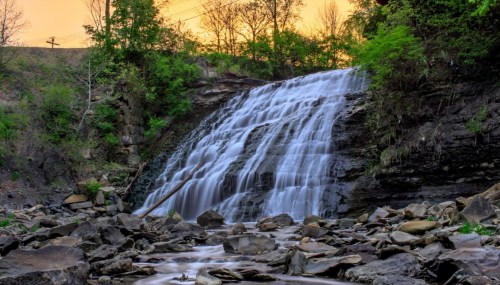 A serene waterfall cascades over rocky terrain, surrounded by lush greenery under a soft sunset sky.