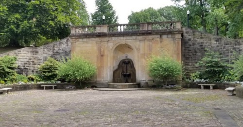 A stone fountain surrounded by greenery and benches in a serene park setting.