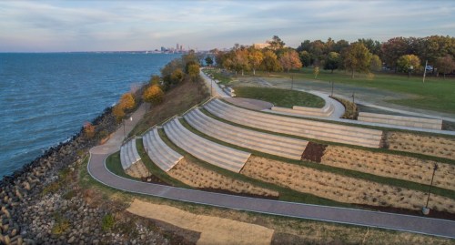Aerial view of a park by the water, featuring landscaped terraces and a path along the shoreline with city skyline in the distance.