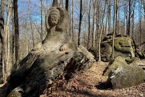 A carved stone figure resembling a seated woman, surrounded by trees and rocky terrain in a forest setting.