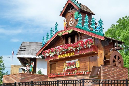 A whimsical wooden building with a clock, flowers, and trees, labeled "Sugarcreek," set against a blue sky.