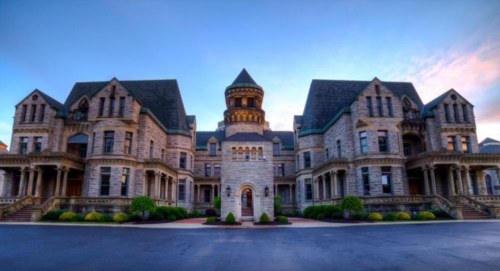 A grand, historic mansion with intricate stonework and a tall tower, set against a colorful sky at dusk.