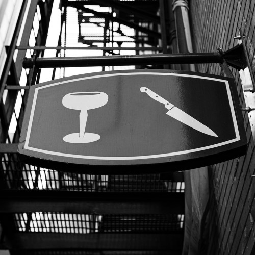 A black and white sign featuring a wine glass and a knife, hanging above a fire escape.