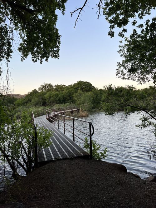 A metal walkway leads over calm water, surrounded by lush greenery and trees under a clear sky.