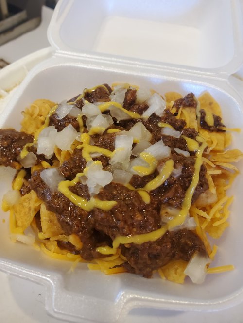 A takeout container filled with nachos topped with chili, shredded cheese, diced onions, and mustard.