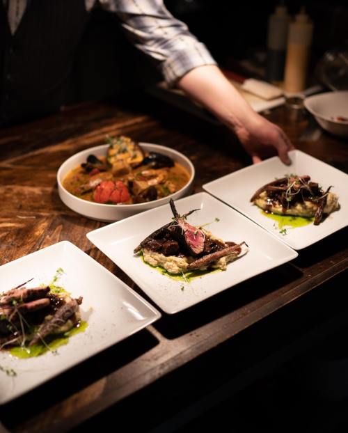 A chef serves gourmet dishes on white plates, featuring meat, vegetables, and garnishes, in a dimly lit restaurant.