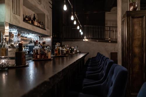 A dimly lit bar with a long counter, lined with bottles and plush blue chairs, creating a cozy atmosphere.
