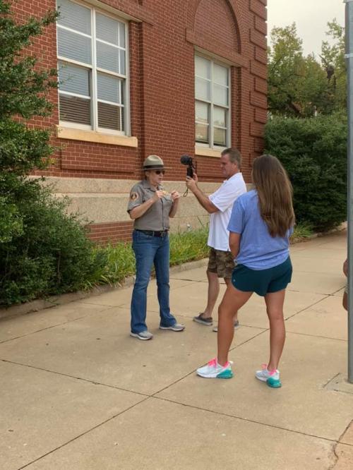 A park ranger speaks to a man filming while a woman listens nearby, outside a brick building.