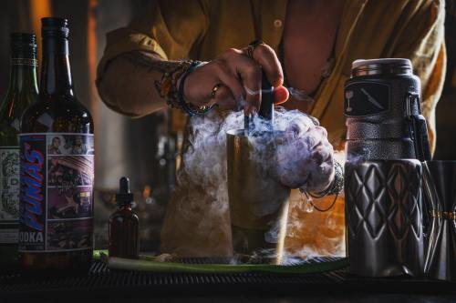 A bartender mixes a cocktail with smoke rising from a shaker, surrounded by various bottles and tools.