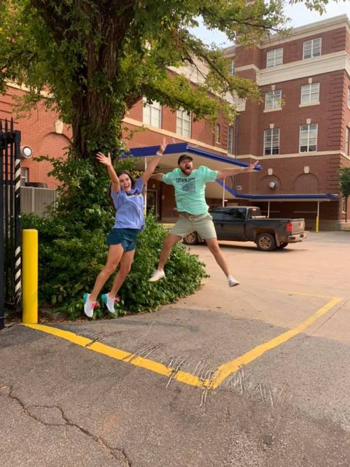 Two people jump joyfully in front of a building, surrounded by greenery and a parked truck.
