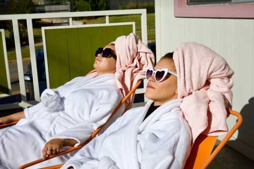 Two women in white robes and sunglasses relax on lounge chairs, with towels wrapped around their heads, enjoying the sun.