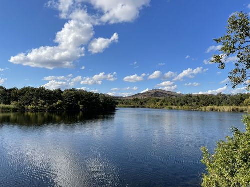 A serene lake surrounded by trees, with a clear blue sky and fluffy clouds reflecting on the water's surface.