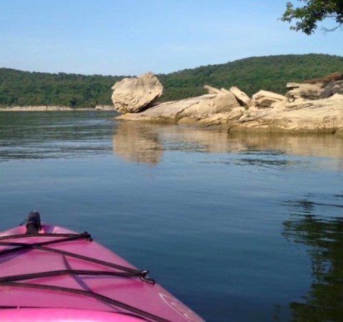 A pink kayak on calm water, with rocky shores and green hills in the background under a clear blue sky.