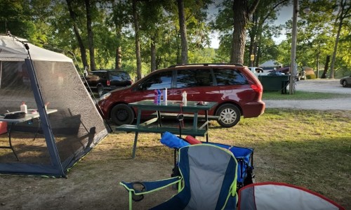 A red minivan parked near a campsite with a picnic table and camping chairs in a wooded area.