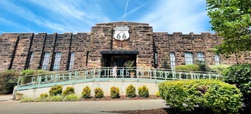 Historic stone building with a Route 66 sign, surrounded by greenery and a blue sky.