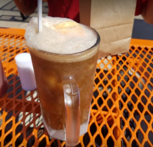A frothy root beer float in a glass mug on an orange table, with a straw and a paper bag in the background.