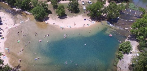 Aerial view of a sandy beach by a clear river, with people swimming and relaxing under trees on a sunny day.