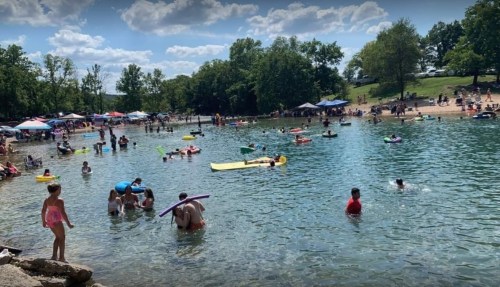 A busy beach scene with people swimming, lounging on floats, and enjoying the sun under blue skies and scattered clouds.