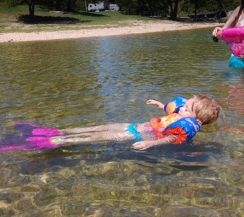 A child floats in shallow water, wearing colorful floaties and swim gear, enjoying a sunny day by the shore.