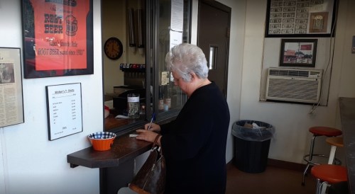 A woman with gray hair writes at a counter in a casual diner, with a menu and decor visible in the background.