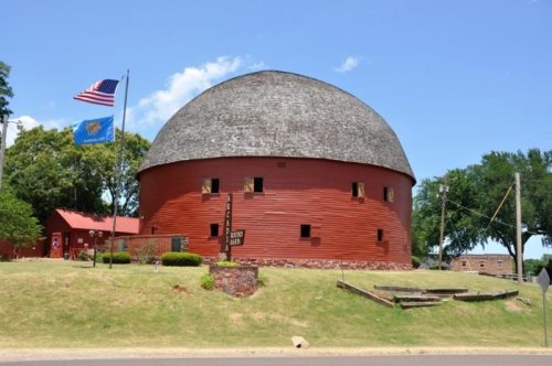A red, round barn with a domed roof, surrounded by green grass and trees, under a clear blue sky. An American flag flies nearby.