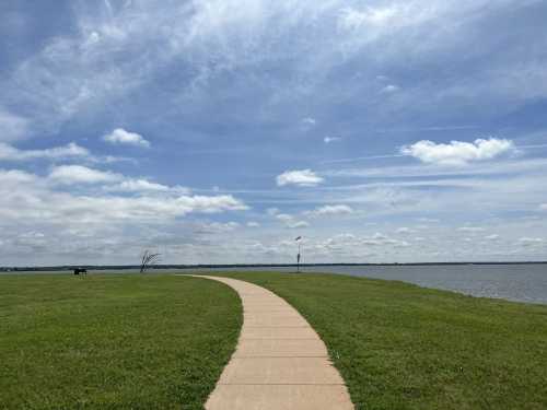 A winding path leads through green grass by a calm lake under a partly cloudy sky.