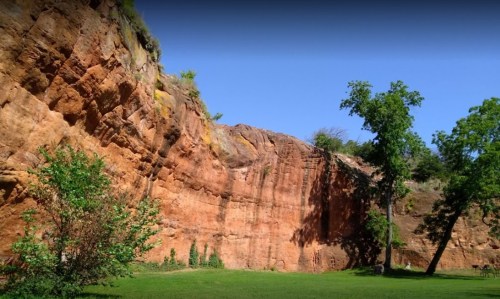 A rocky cliff with reddish hues, surrounded by green grass and trees under a clear blue sky.