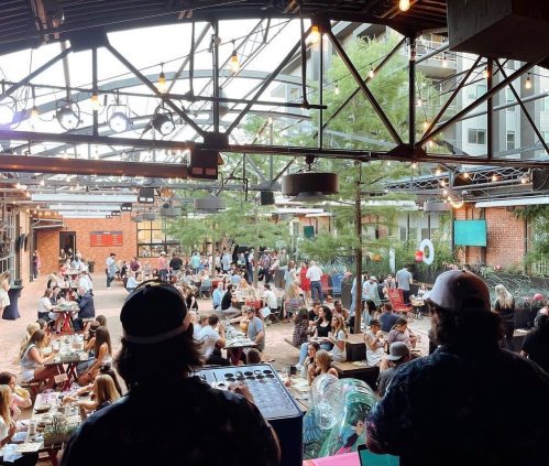 A lively outdoor venue filled with people dining and socializing under string lights and greenery.