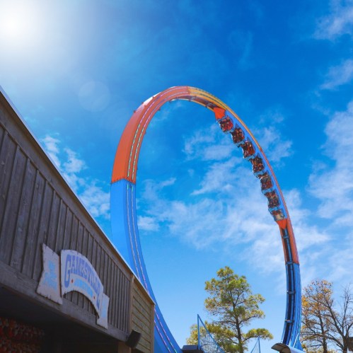 A colorful amusement park ride arcs against a bright blue sky with scattered clouds and trees in the background.