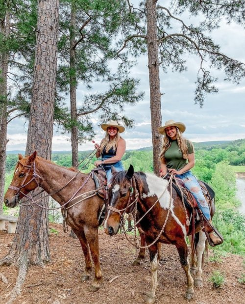 Two women on horseback in a forested area, wearing cowboy hats and casual attire, smiling at the camera.