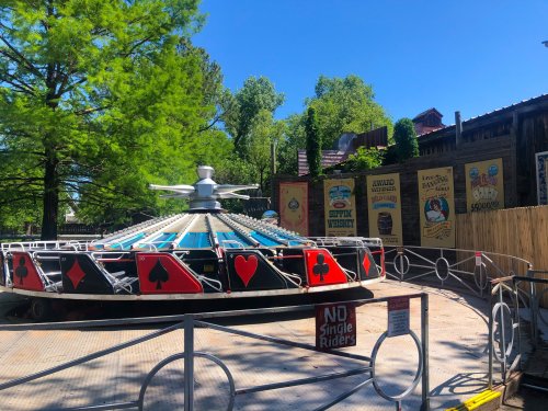A spinning amusement ride with playing card designs, surrounded by trees and colorful signage in a sunny park.