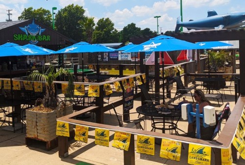 Outdoor dining area at Shark Bar with blue umbrellas, tables, and banners, featuring a relaxed, tropical atmosphere.