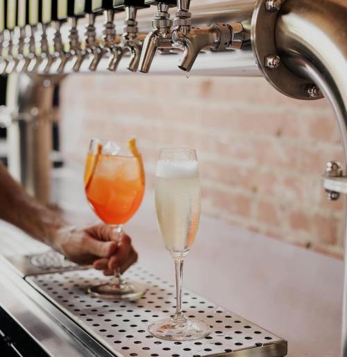 A hand holds a colorful cocktail and a champagne flute in front of a sleek bar tap setup.