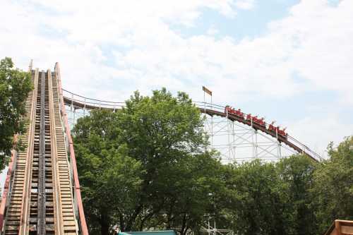 A roller coaster climbs steeply, with riders at the top, surrounded by trees and a cloudy sky.