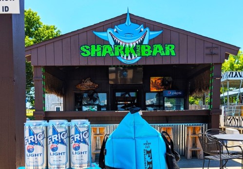 A beach bar named "Shark Bar" with a shark logo, featuring drinks and outdoor seating.