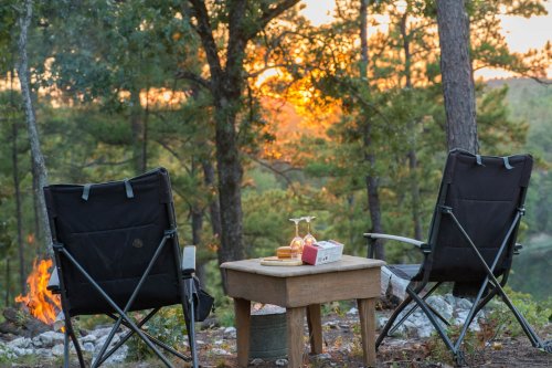 Two camping chairs facing a small table with snacks, set against a sunset backdrop in a forested area.