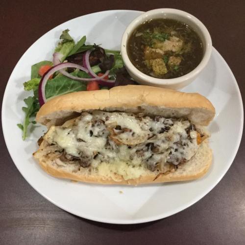 A sandwich filled with melted cheese, onions, and meat, served with a side salad and a bowl of soup.