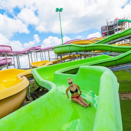 A person slides down a green water slide at a water park, surrounded by colorful slides and a clear blue sky.