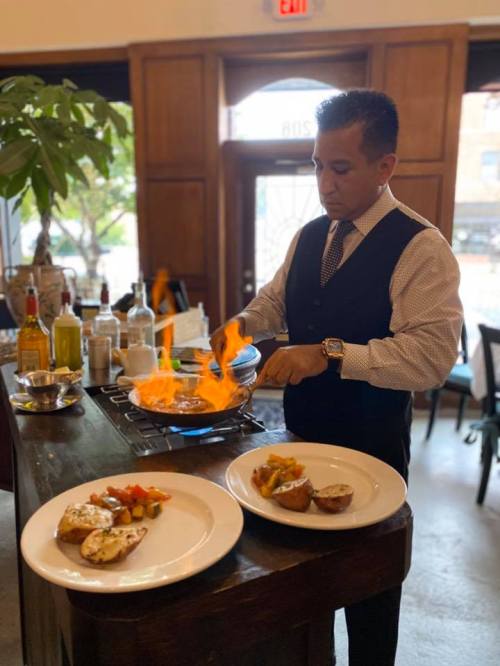 A chef in a vest cooks with flames in a pan, with two plated dishes on the table in a restaurant setting.