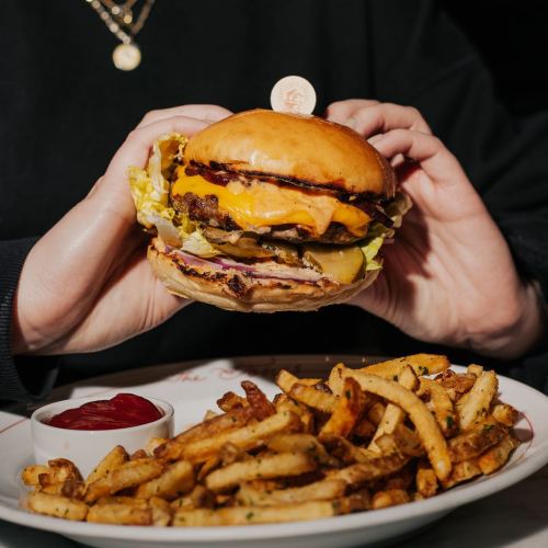 A person holds a cheeseburger with lettuce and pickles, accompanied by a plate of golden fries and a side of ketchup.