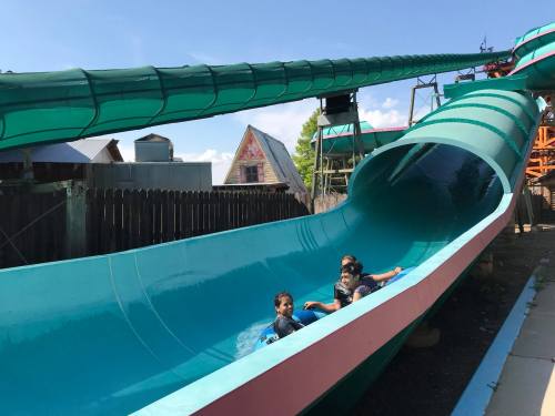 Two children enjoy a water slide on a sunny day, surrounded by green and blue structures.