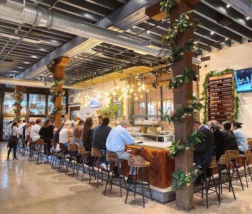 A bustling bar with patrons seated at a long counter, surrounded by greenery and warm lighting in a modern space.