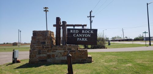 Sign for Red Rock Canyon Park, featuring a stone base and wooden structure, set in a grassy area with utility poles nearby.