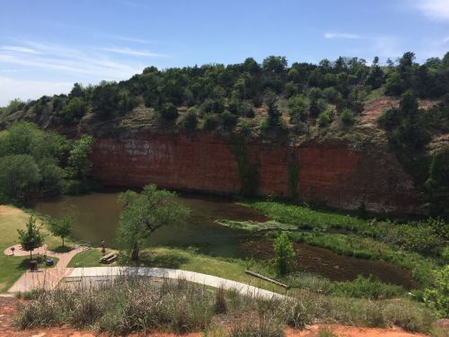 A serene landscape featuring a calm pond surrounded by lush greenery and a steep, red rock cliff in the background.