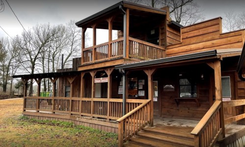 A rustic wooden building with multiple balconies and a welcoming entrance, surrounded by trees and a grassy area.