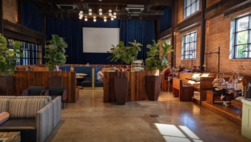 A spacious restaurant with wooden decor, plants, and diners enjoying meals in a cozy, modern atmosphere.