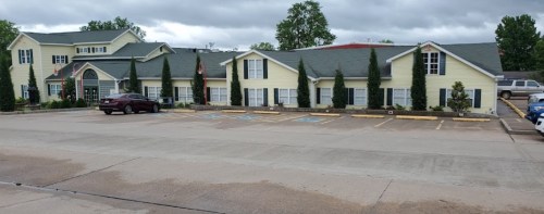 A yellow two-story building with a green roof, surrounded by trees, and a parking lot with several cars.