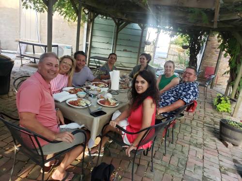 A group of people enjoying a meal together outdoors under a shaded pergola, surrounded by greenery.